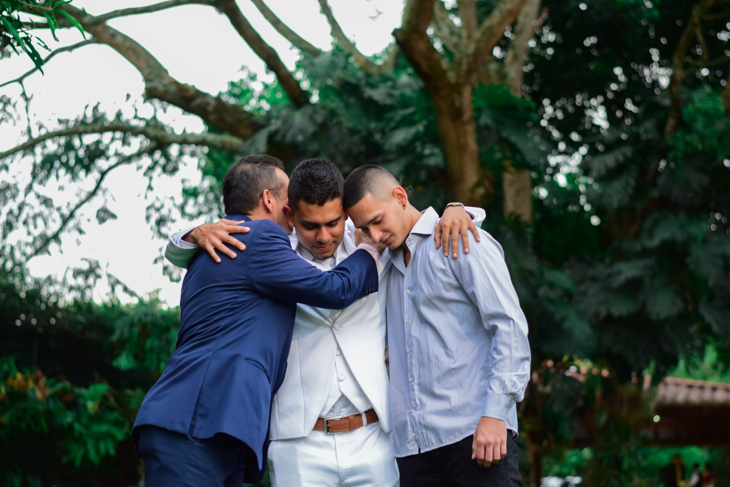 Three men embrace outdoors, symbolizing friendship and support in a natural setting.